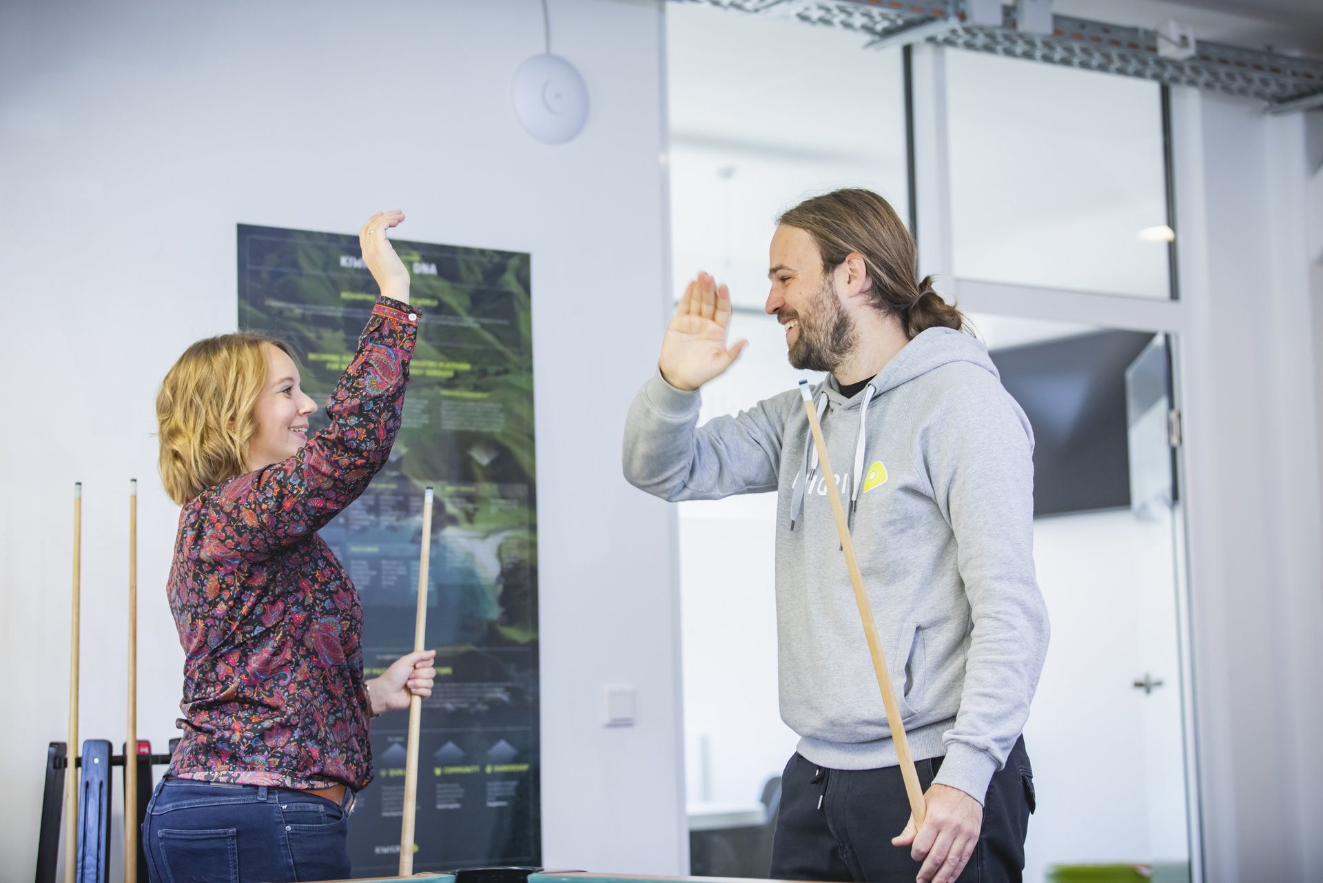 Woman and man in Kiwigrid hoodie play pool in the office and high-five each other.